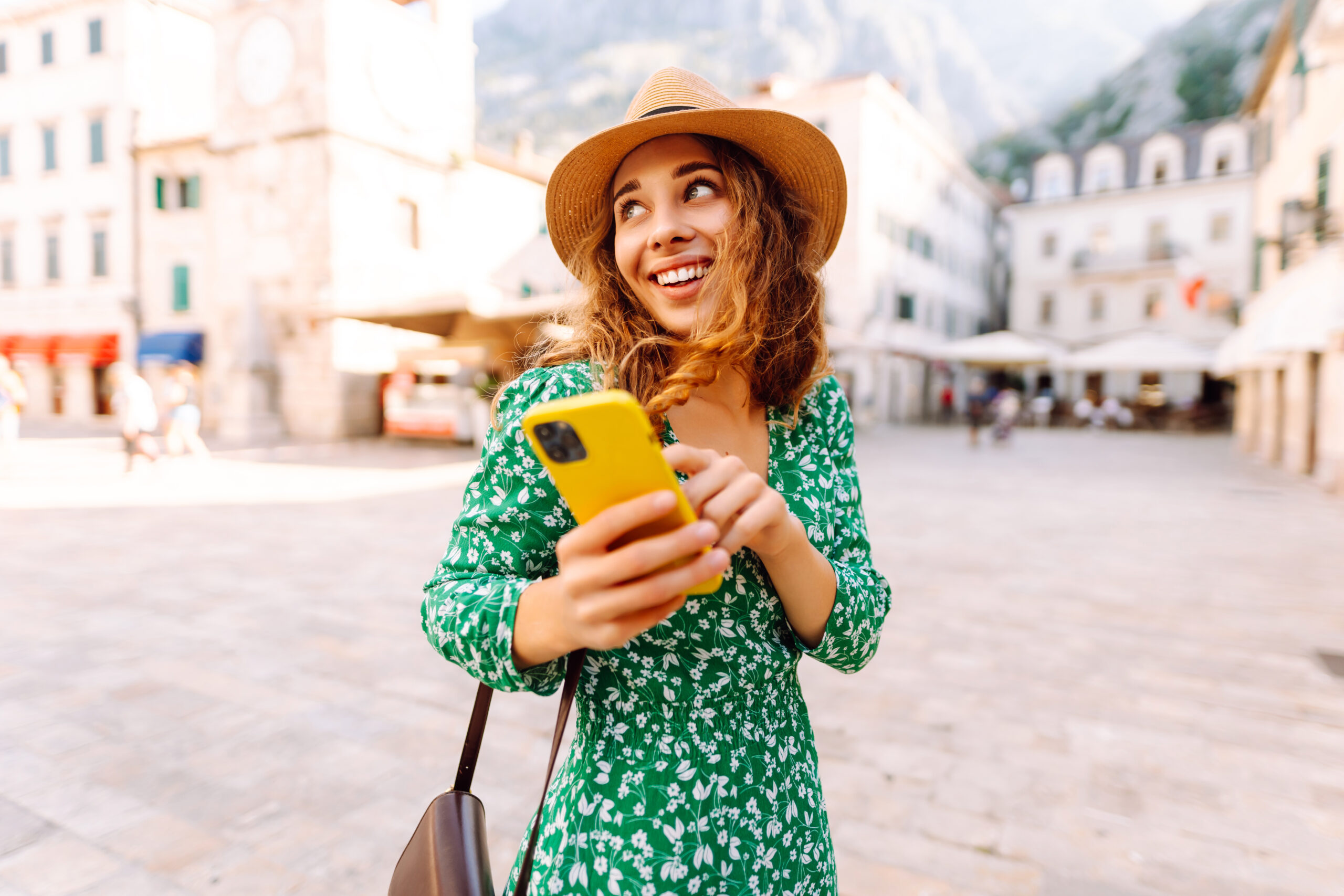 Young woman in a green dress smiles while using her yellow smartphone in a quaint European plaza on a sunny day. Travel, tourism, technology, blogging, communication concept.