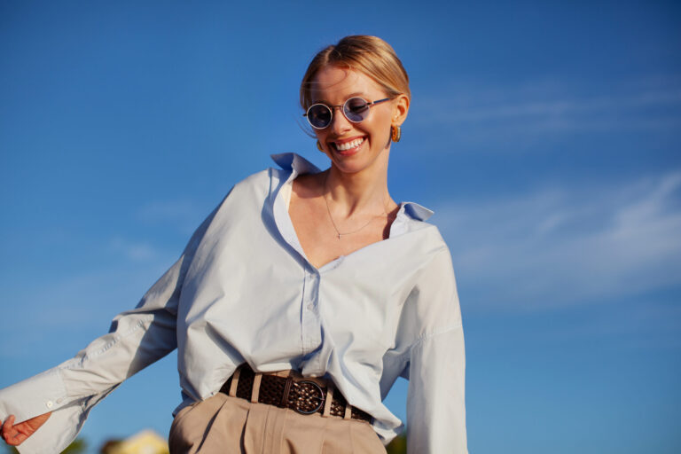 Contemporary happy woman in fancy clothes oversized shirt and sunglasses, joyfully smiling against blue sky. Trendy, happy, and stylish summer vibes