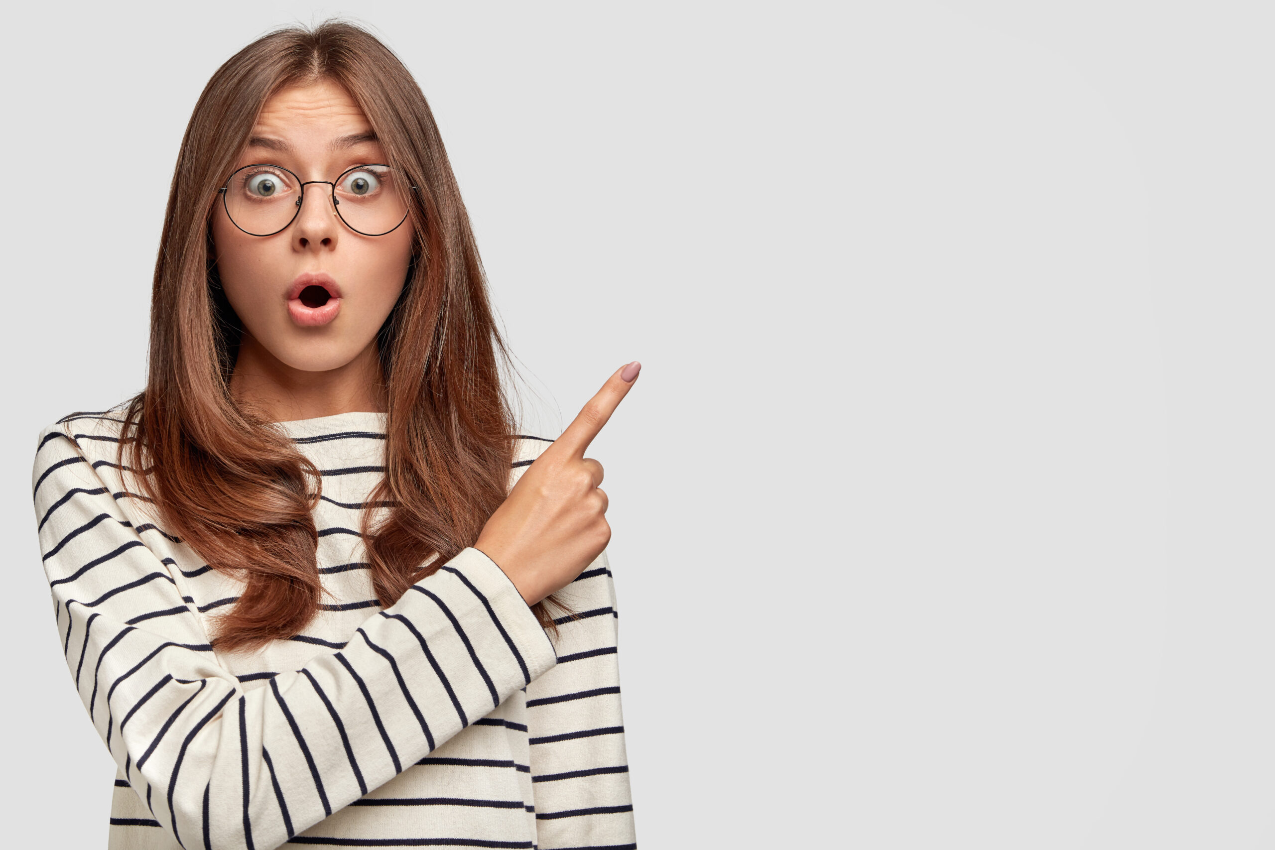 Horizontal shot of beautiful young Caucasian female with surprised expression, points at upper right corner, dressed in striped sweater, wears round spectacles, isolated over white background