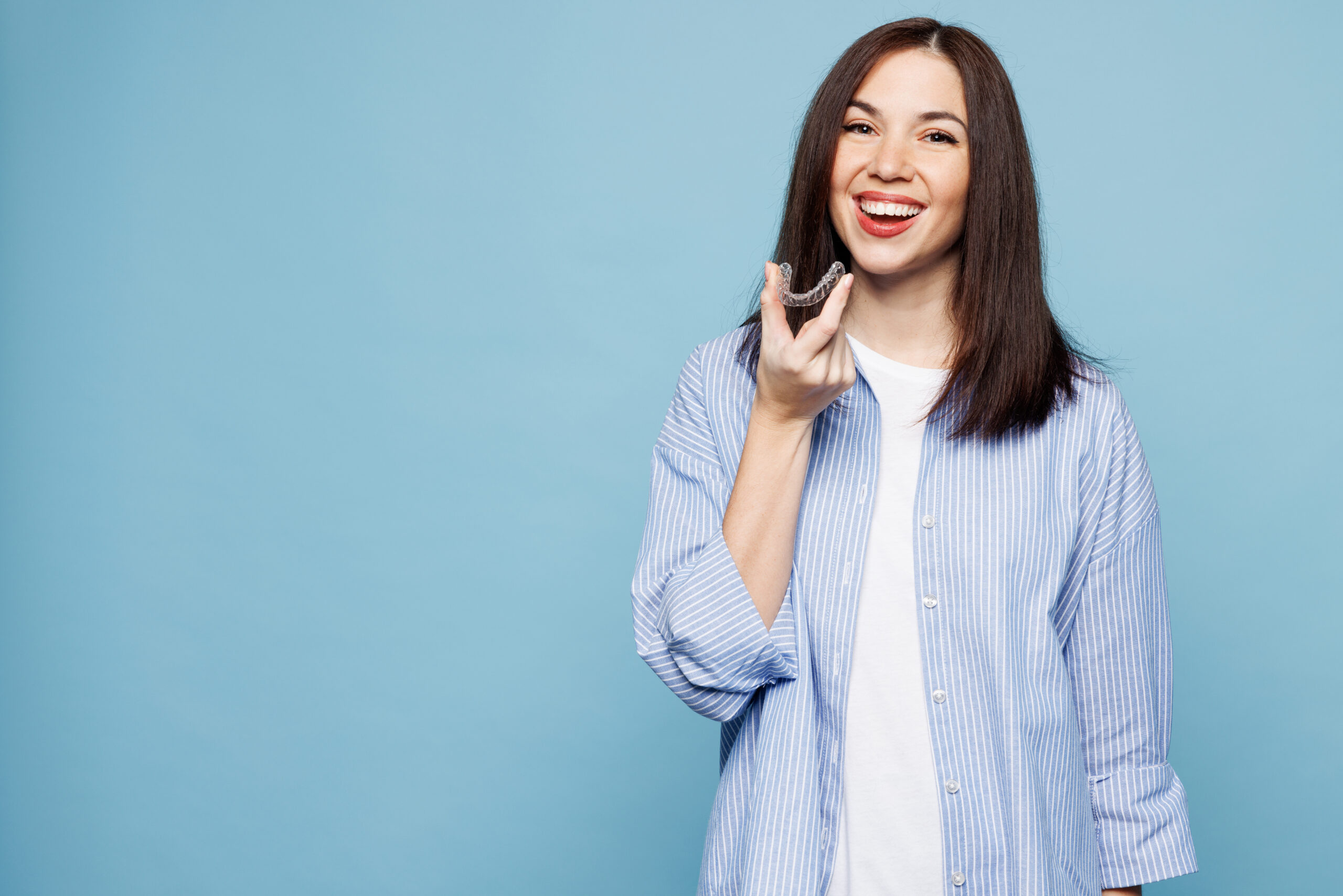 Young smiling woman she wearing shirt casual clothes hold in hand invisible transparent aligners, invisalign dental braces isolated on plain pastel light blue cyan background studio Lifestyle concept