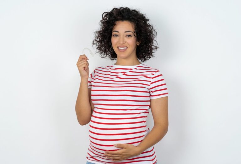 young pregnant woman wearing striped t-shirt over white background holding an invisible braces aligner, recommending this new treatment. Dental healthcare concept.