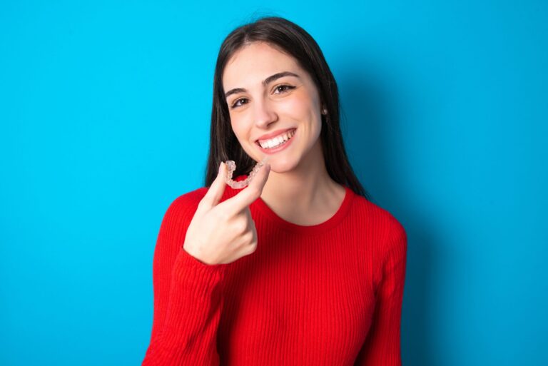 young brunette girl wearing red T-shirt against blue wall holding an invisible aligner ready to use it. Dental healthcare and confidence concept.