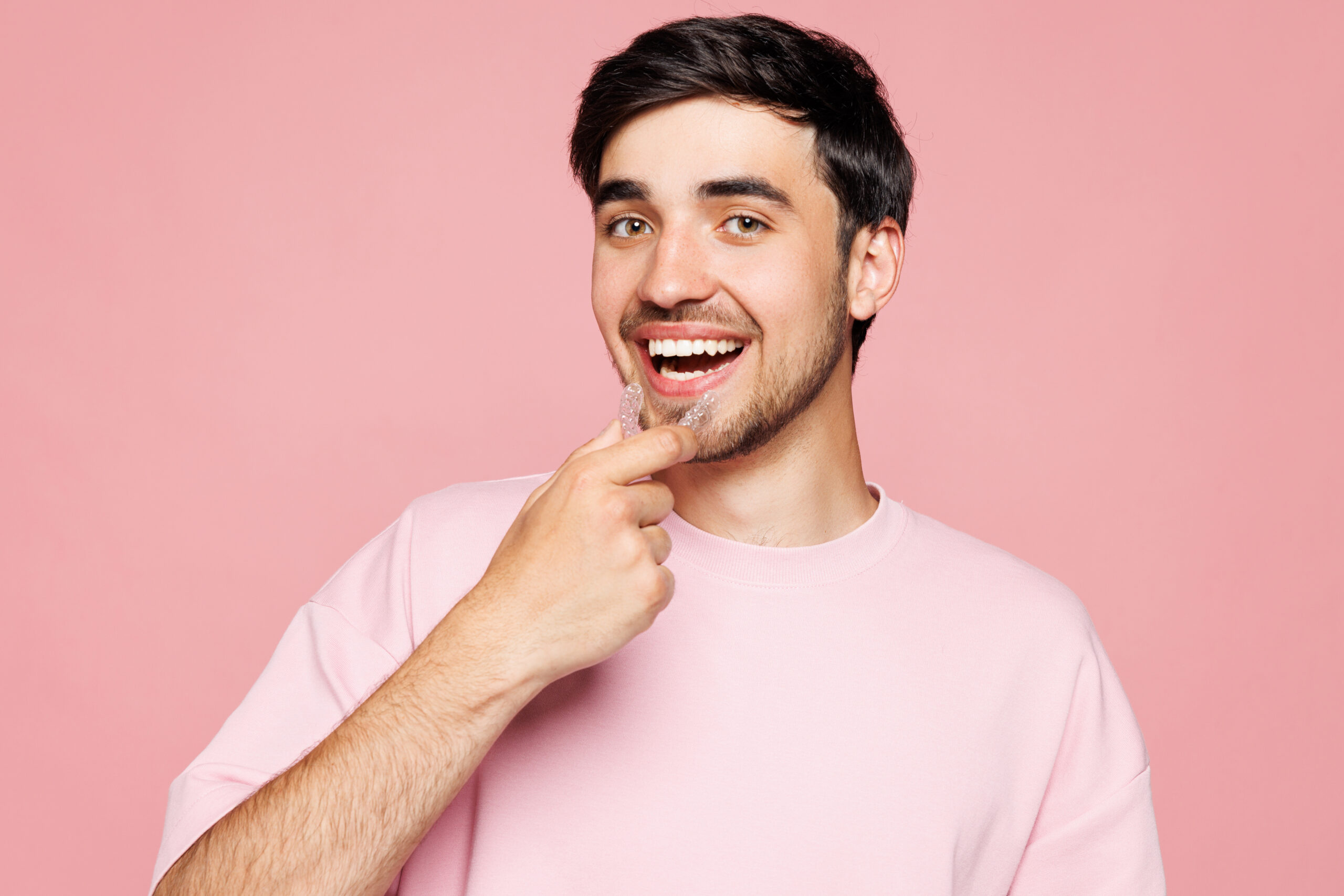 Close up young smiling man wear t-shirt casual clothes hold in hand invisible transparent aligners, silicone invisalign dental braces isolated on plain pastel light pink background. Lifestyle concept.