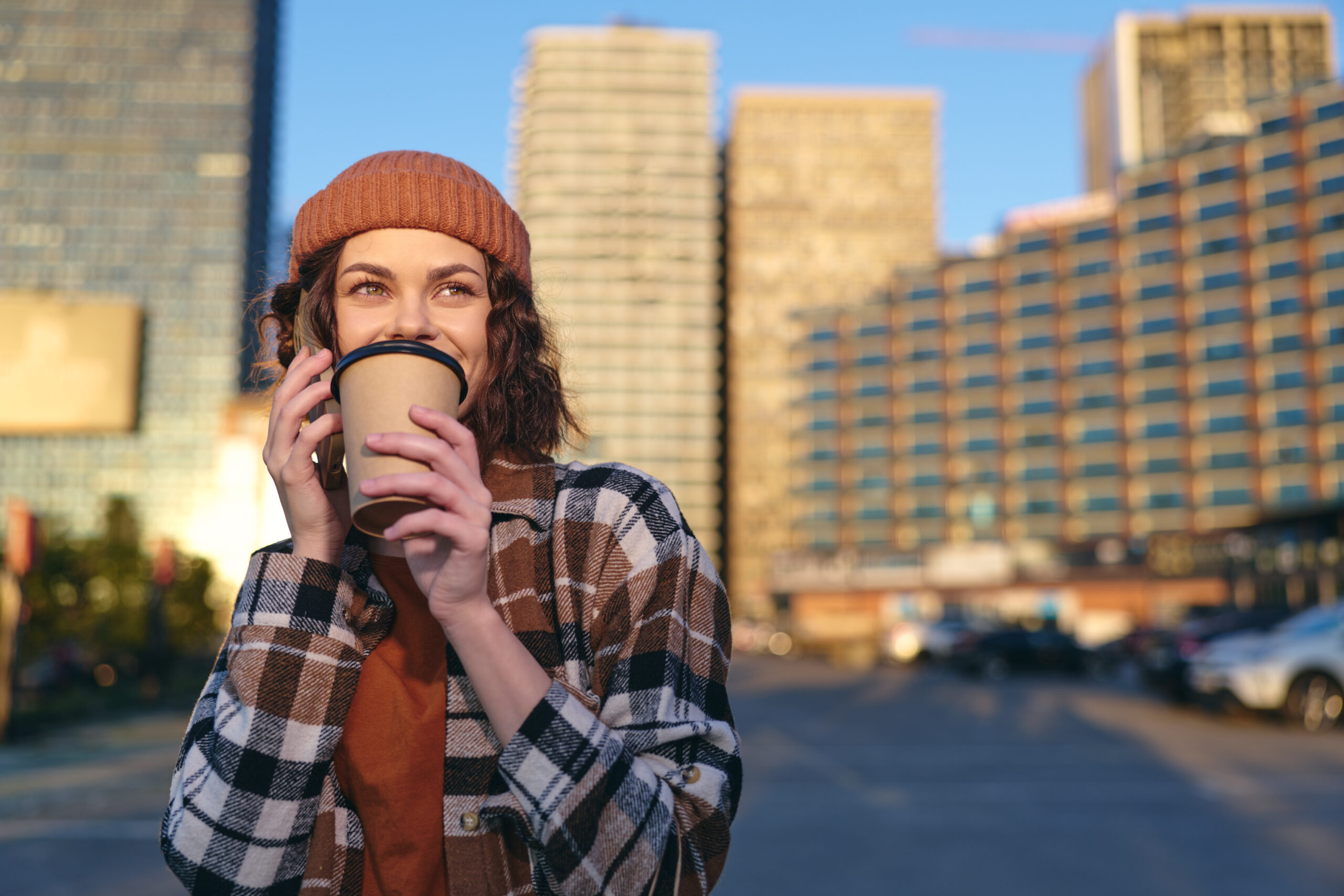 Woman holding coffee and phone with a smile in urban street at golden hour, candid lifestyle portrait in beanie, authenticity and mindful living with emotional storytelling glow.